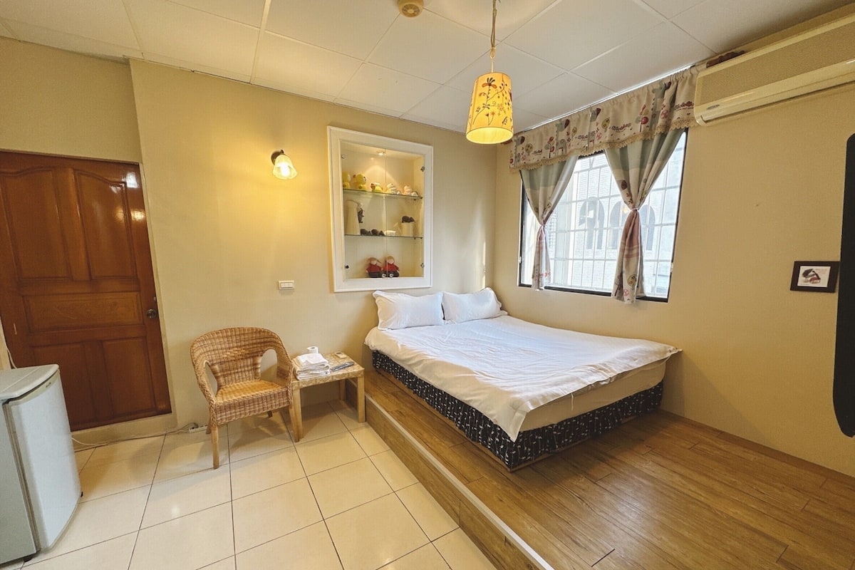 A bedroom with a bed, a chair, a small table, a mirror, and a window with curtains.