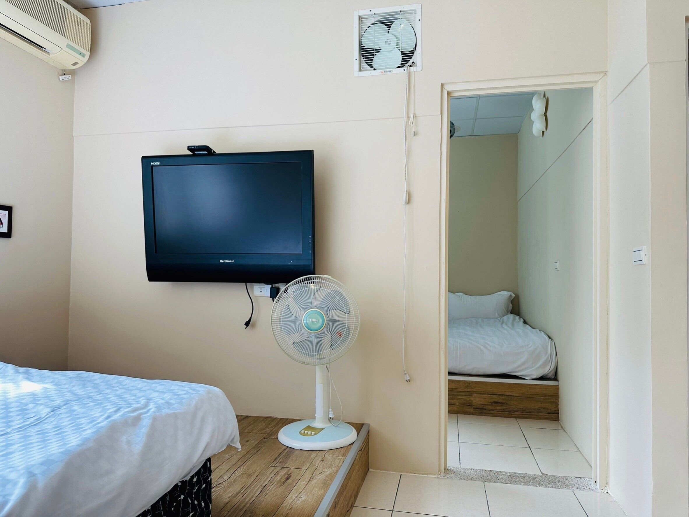 A bedroom with a bed, a wall-mounted TV, a standing fan, and a door leading to another room.