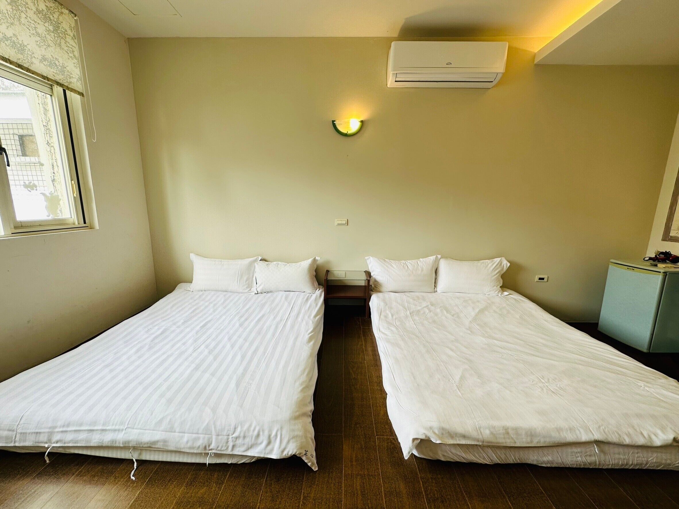 A room with two single beds, a window with a view, an air conditioner, and a small cabinet.
