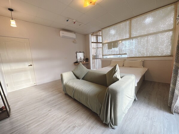 A room with a bed, a sofa, a window with lace curtains, a wall-mounted air conditioner, and a door.