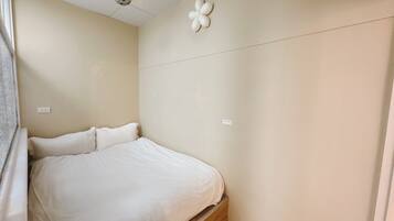 A small, neatly made bed in a room with a plain wall and a ceiling-mounted light fixture.