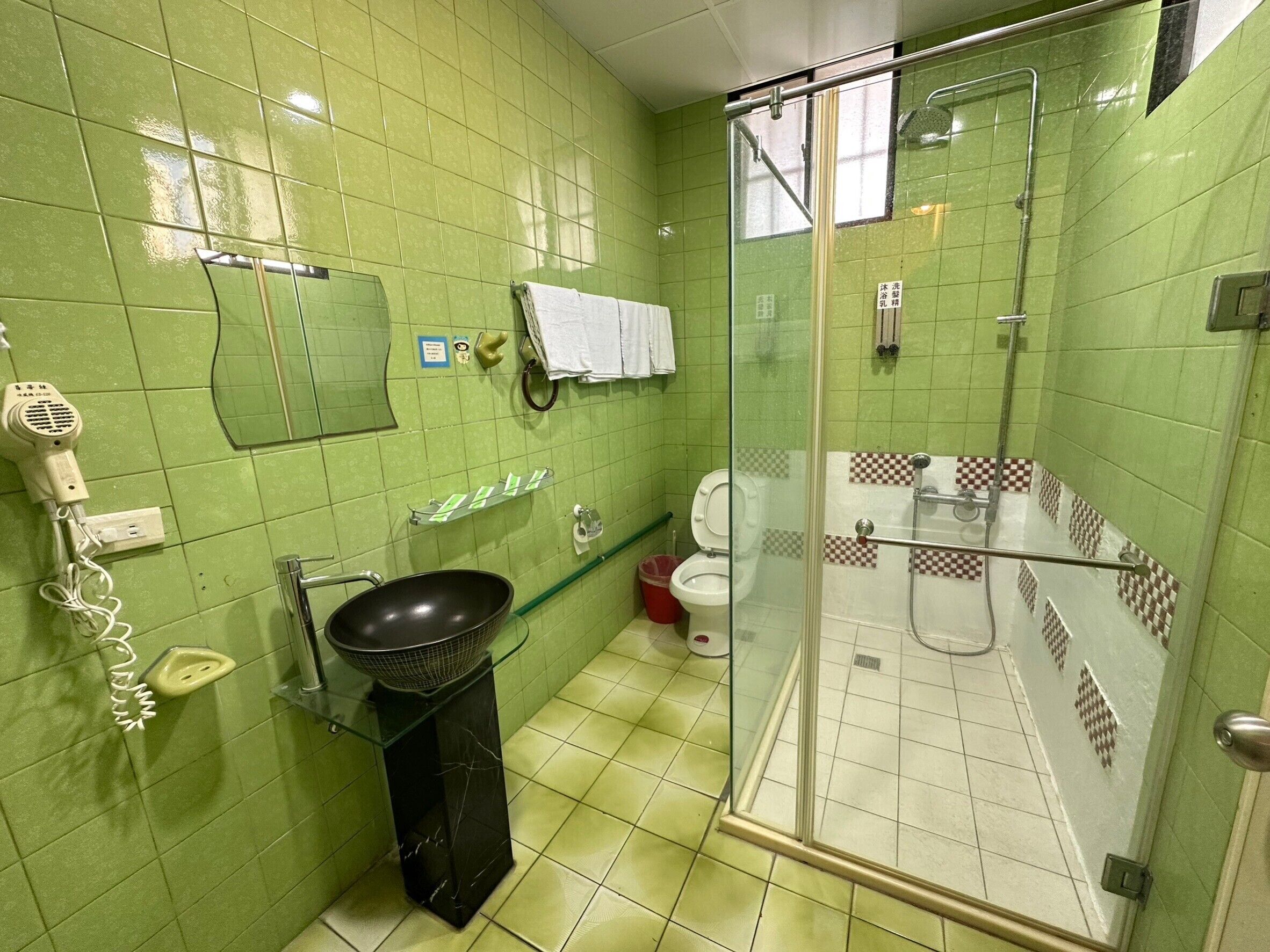 A bathroom with green tiles, a glass-enclosed shower, a sink with a black basin, a white toilet, and a hairdryer on the wall.