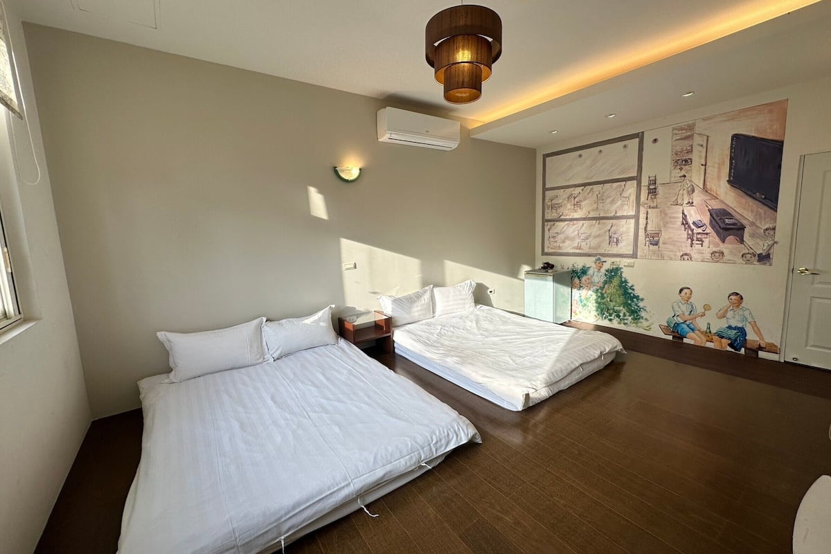 A bedroom with two beds, a wooden floor, a ceiling light, and a wall mural.