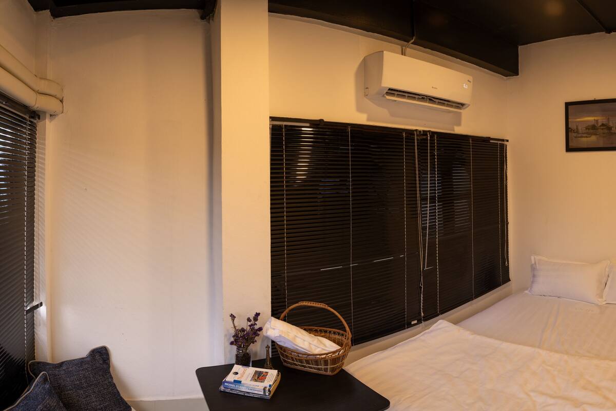 Una habitación de hotel con una cama, una mesita con un libro y una cesta, aire acondicionado en la pared y una ventana con persianas.