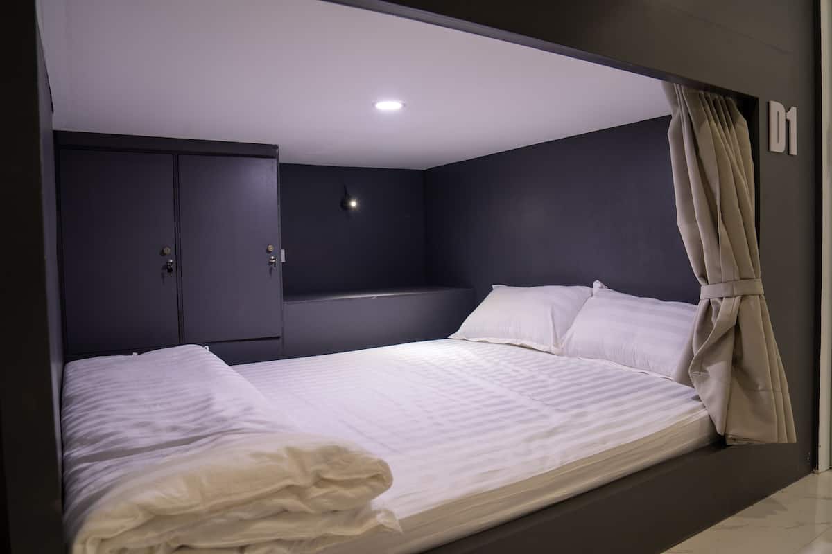 Una cama individual con ropa de cama blanca, una pared oscura, una lámpara en el techo y una puerta con el símbolo del número.