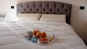 Ein ordentlich bezogenes Bett mit grauem, gepolstertem Kopfteil, weißen Kissen und einem Tablett mit einer Tasse Tee, zwei Muffins und einer kleinen Snacks-Verpackung.