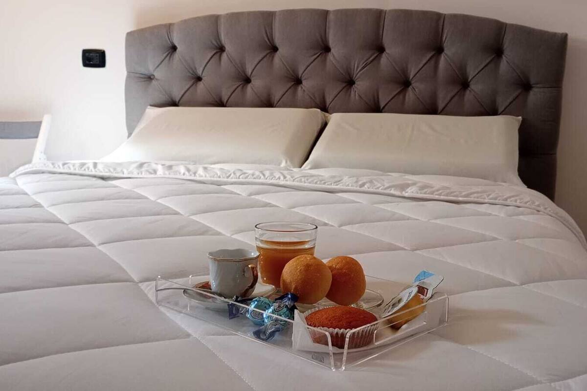 Un lit bien fait, avec une tête de lit capitonnée grise, des oreillers blancs et un plateau contenant un verre de thé, deux muffins et une petite boîte de collations.