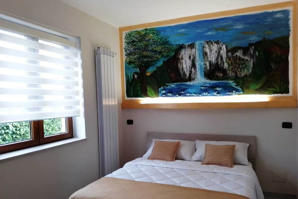 Une chambre à coucher avec un lit, une fenêtre avec des stores et un tableau représentant une cascade accroché au mur.