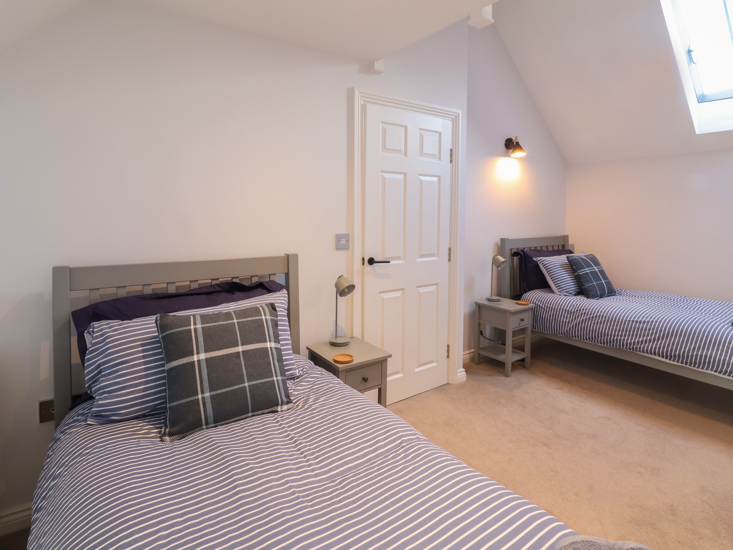 A bedroom with two beds, a door, a nightstand, a lamp, and a skylight.