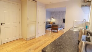 A modern kitchen with wooden flooring, a black countertop, and stainless steel appliances. There is a grey sofa and a small table in the living area, and a white wardrobe with silver handles. - Spring Rose (Bedale)