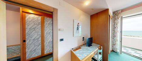A hotel room with a wooden desk, a flat-screen TV, a framed picture on the wall, and a view of the sea through the window.
