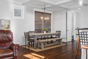 Un comedor con mesa de madera, sillas y banco. Hay una ventana con persianas, una lámpara colgante y un cuadro en la pared.