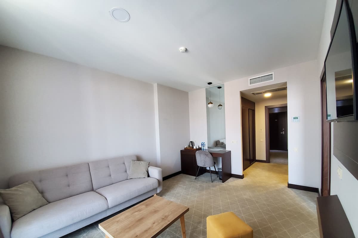 A modern hotel room with a sofa, a wooden coffee table, a small ottoman, a desk with a chair, and a large mirror.