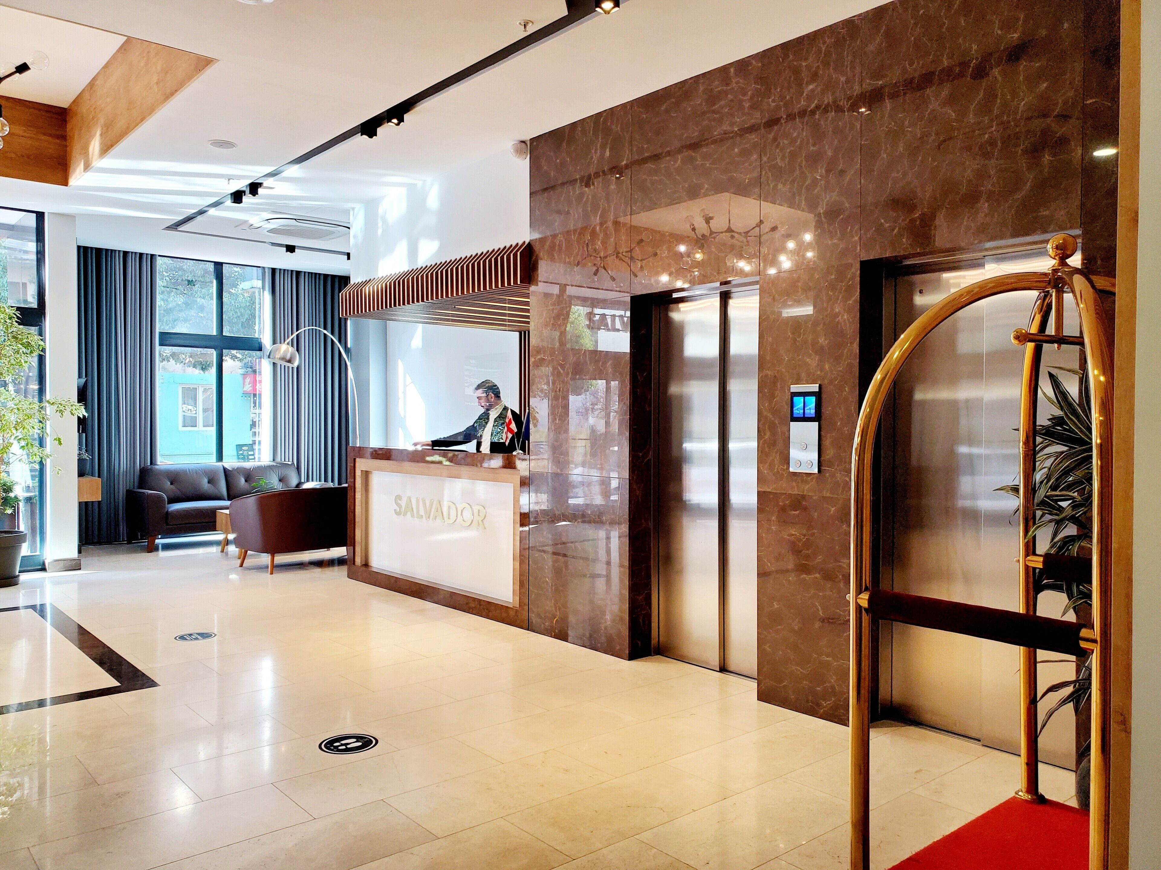 Un hall d’hôtel avec un comptoir de réception portant l’inscription « SALVADOR », un espace d’accueil avec un canapé et des fauteuils, un grand miroir et un tapis rouge menant à un ascenseur.