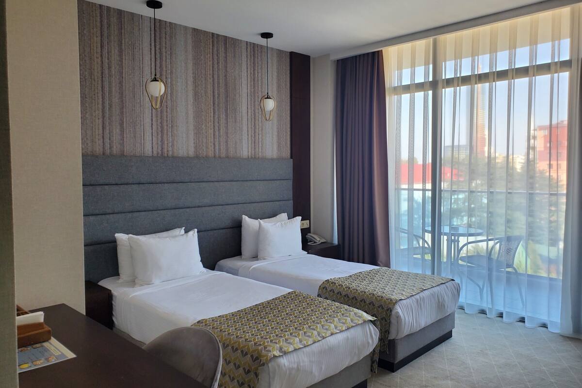 A hotel room with two beds, a large window with curtains, a headboard, and a small table with a chair.
