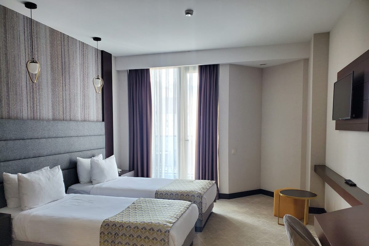 A hotel room with two beds, a large window with curtains, a flat-screen TV, and a small round table.