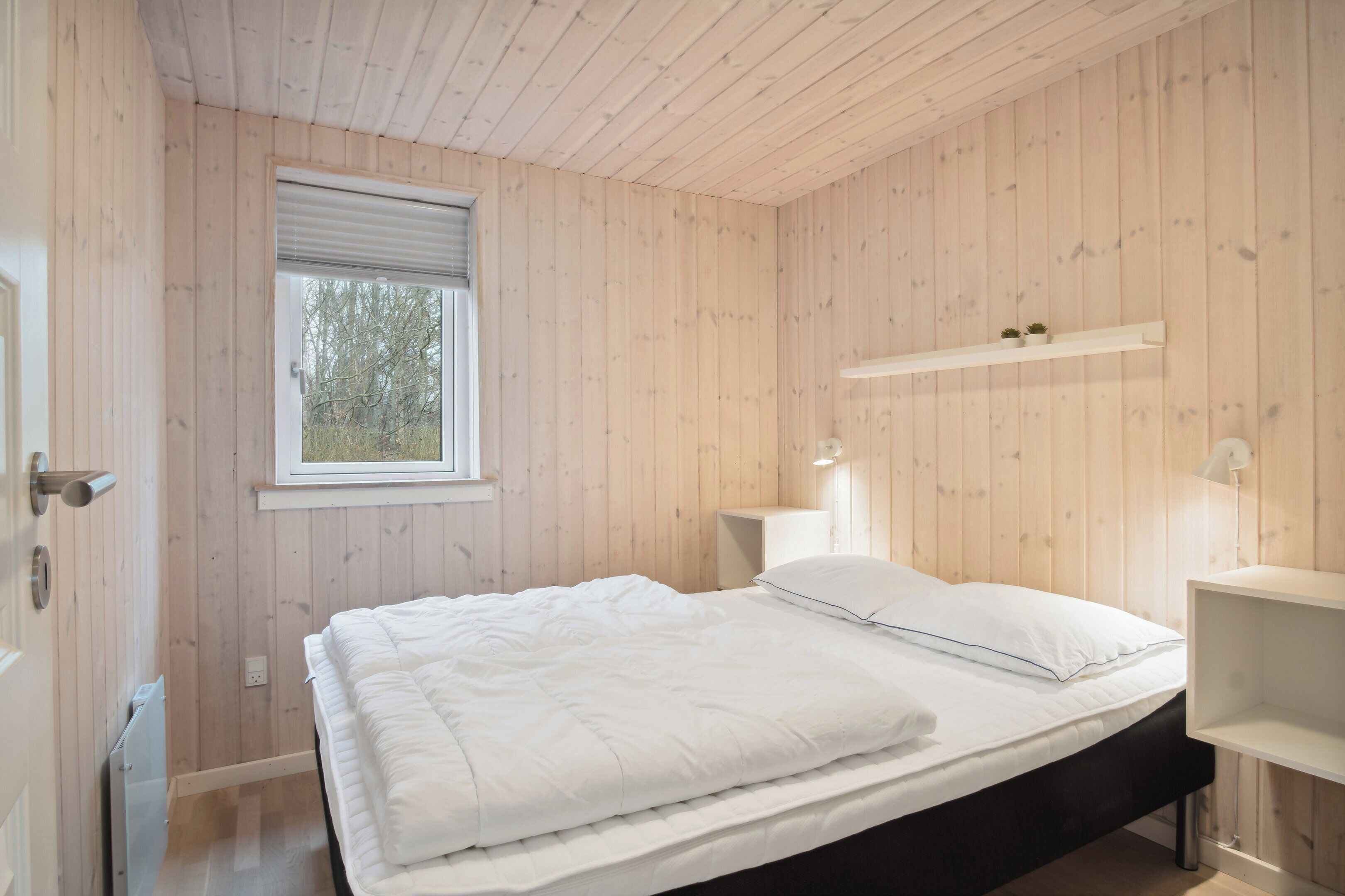 Een kamer met houten wanden, een bed met wit beddengoed, een raam en een plankje met twee kleine planten.