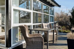 A covered outdoor deck with wicker furniture and a table, large windows, and a view of trees and buildings.