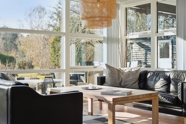 A modern living room with a large window, a wooden coffee table, a black leather sofa, and a wicker hanging light fixture.
