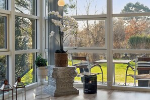 A room with large windows, a stone pedestal with a plant, a metal chair, and a table.