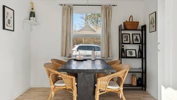 A dining area with a dark wooden table, wicker chairs, a white car visible through the window, and a shelf with bottles and pictures.