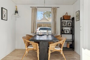 A dining area with a dark wooden table, wicker chairs, a white car visible through the window, and a shelf with bottles and pictures.