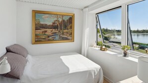 A bedroom with a bed, a painting, a window with a view of a lake, and a bedside table with a lamp. - Lovely beach house with panoramic view (Martofte)