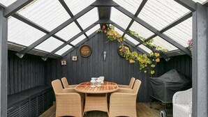 A covered patio with a round table and wicker chairs, a grill, and a wall-mounted sign. - Renovated summer house incl. cleaning and linen (Juelsminde)