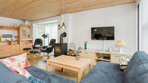 A modern living room with a sofa, a wooden coffee table, a TV, a fireplace, and a kitchen area. - Renovated summer house incl. cleaning and linen (Juelsminde)