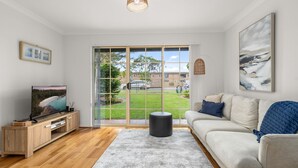 A living room with a large sliding glass door, a flat-screen TV, a wooden cabinet, a grey rug, a beige sofa, and a painting on the wall. - Haw1.29 - Shells by Experience Jervis Bay (Huskisson)