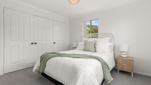 A bedroom with a white bed, a nightstand with a lamp, a window with a view of greenery, and a white wardrobe. - Haw1.29 - Shells by Experience Jervis Bay (Huskisson)