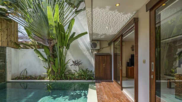 Un área de piscina moderna al aire libre con un deck de madera, una puerta de cristal que da a una habitación y exuberante follaje tropical.