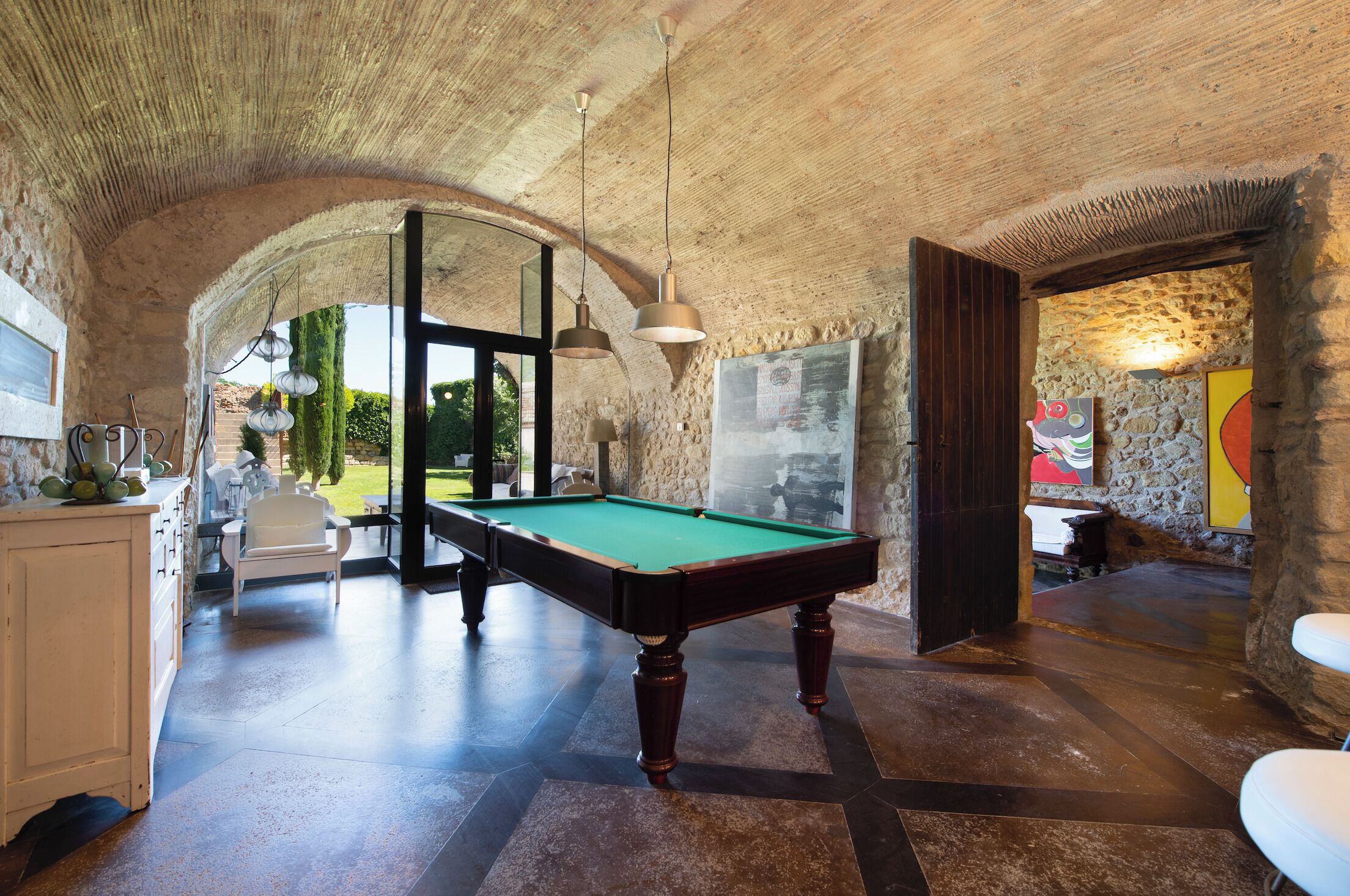 Una mesa de billar en una habitación con paredes de piedra y un techo alto. Hay dos lámparas colgantes, un armario blanco y una puerta que da a otra habitación.