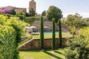 A pool area with stone walls, a view of a historic building, and a well-maintained garden with tall trees.