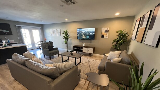A modern living room with a sofa, armchairs, a coffee table, and a TV. There are plants and artwork on the walls.
