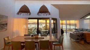 A modern dining area with a wooden table and chairs, two hanging woven light fixtures, and large windows offering a view of a body of water and distant hills.