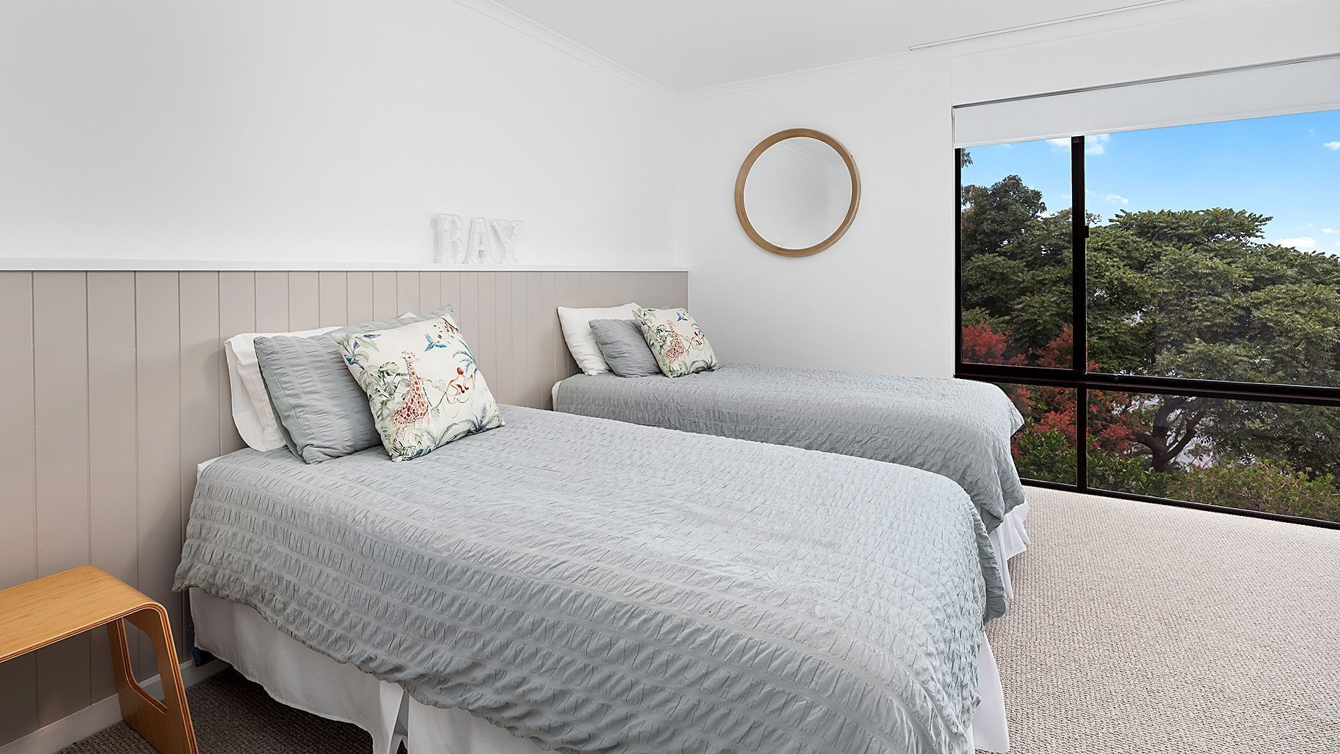 Une chambre avec deux lits, une tête de lit en bois, un grand miroir rond et une grande fenêtre donnant sur les arbres.