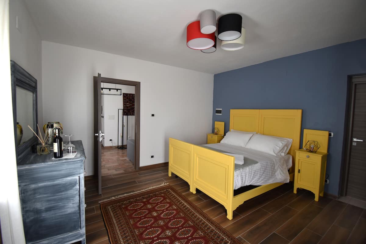 Une chambre avec un lit jaune, une commode en bois, un tapis rouge à motifs et un mur bleu.