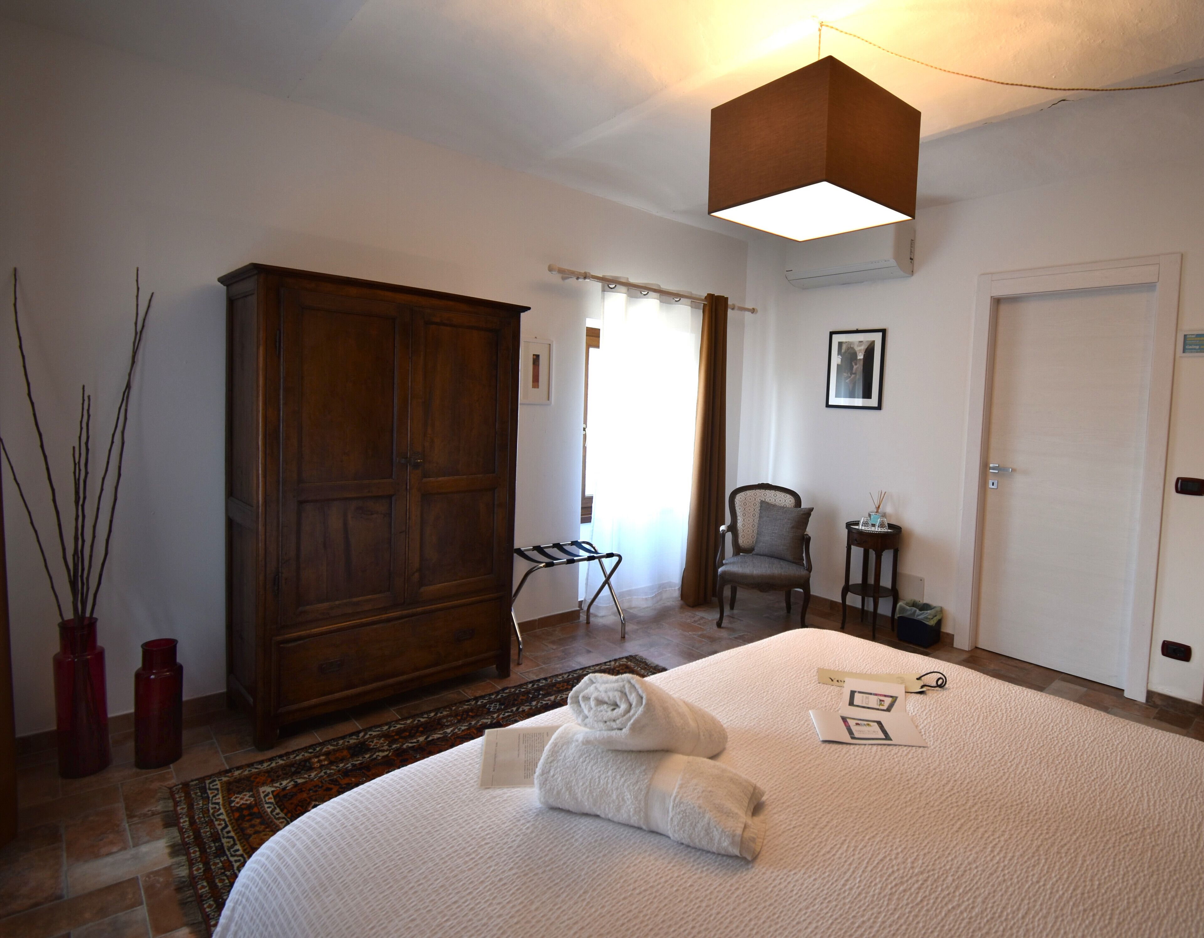 A hotel room with a large wooden wardrobe, a bed with towels, a chair, a small table with a bottle, and a picture on the wall.