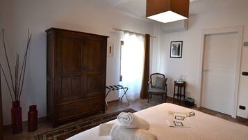 A hotel room with a large wooden wardrobe, a bed with towels, a chair, a small table with a bottle, and a picture on the wall.