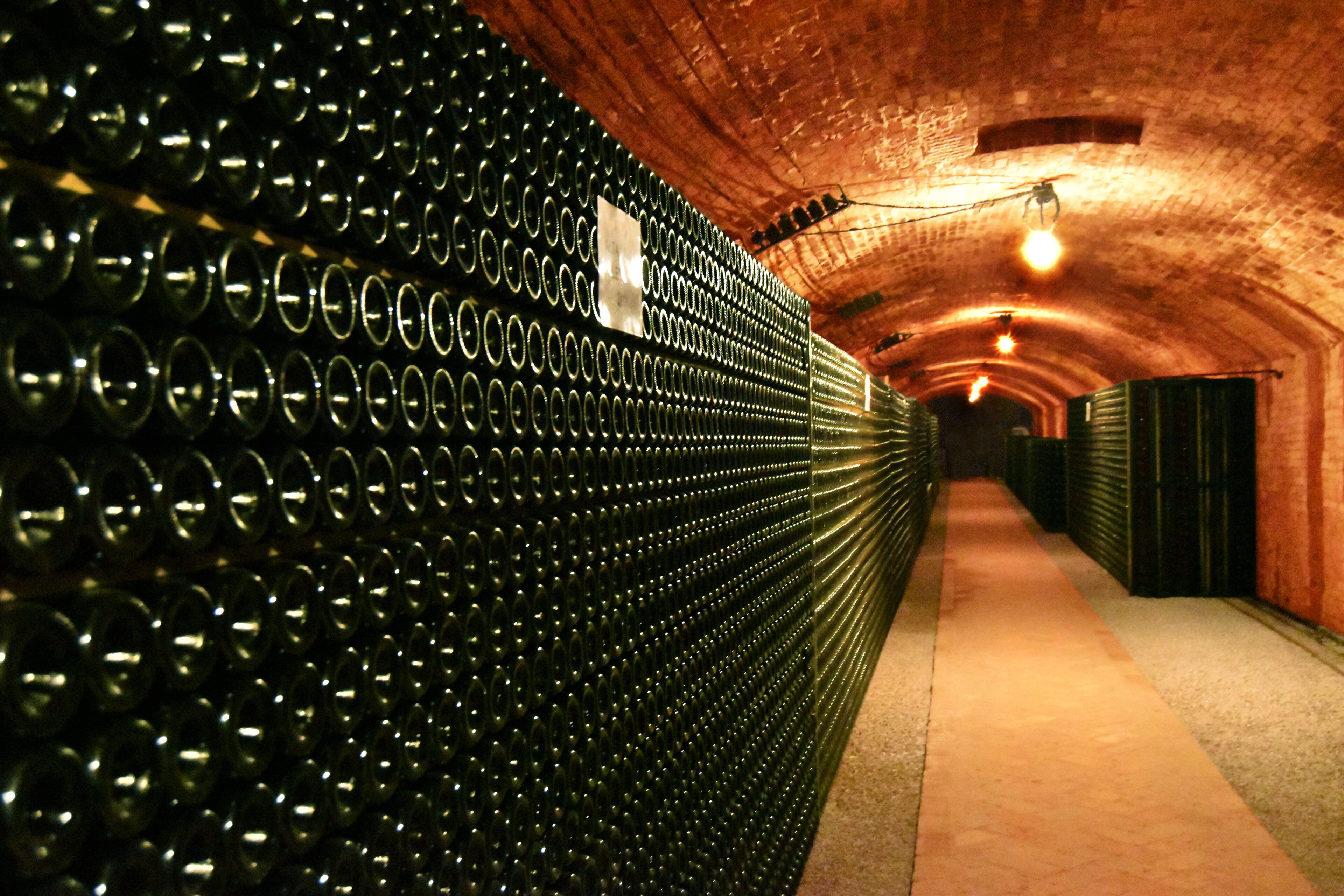Un túnel revestido con hileras de botellas de vino, paredes de ladrillo y una lámpara.