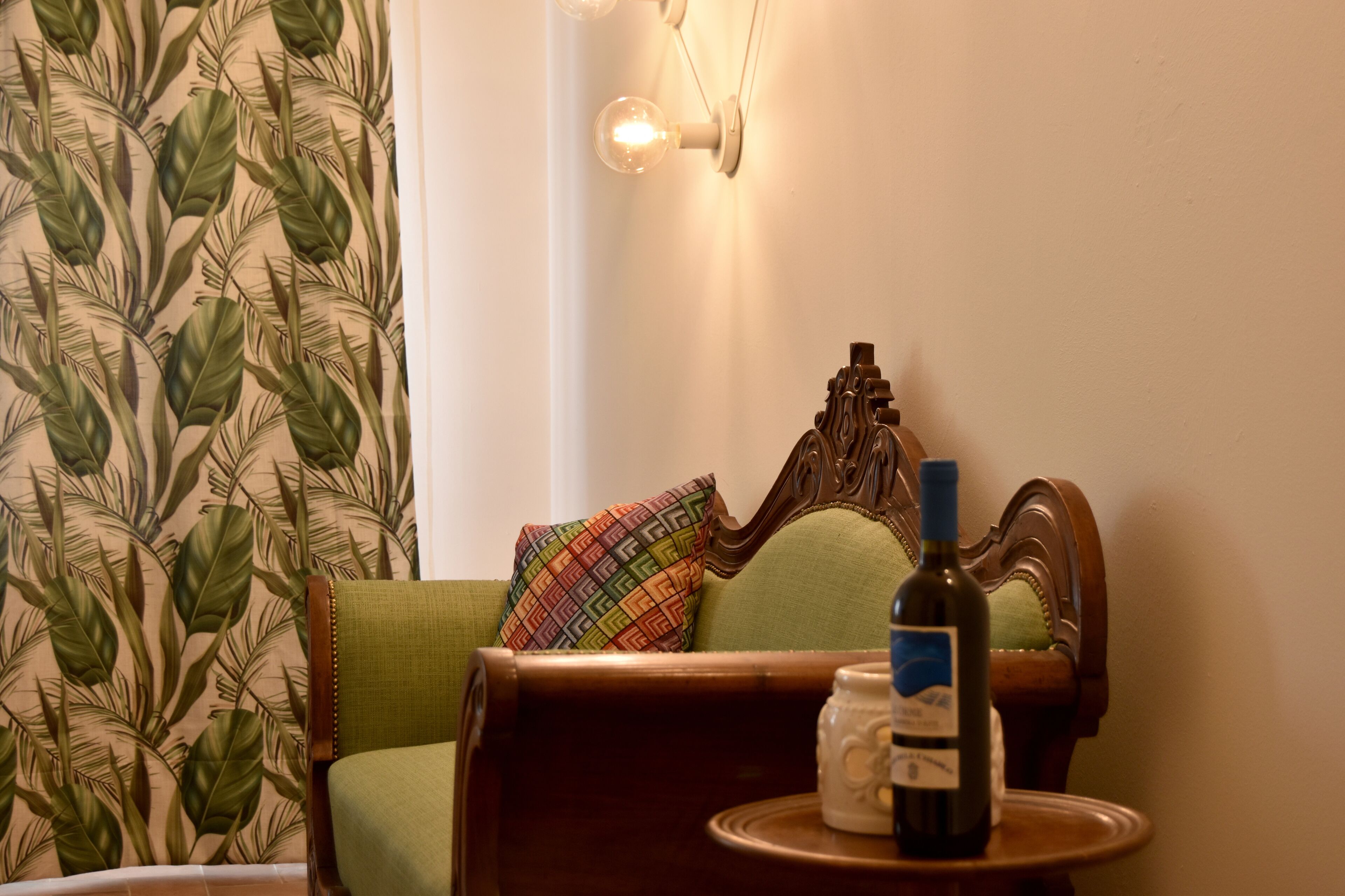 Una habitación acogedora con un sillón verde, un papel tapiz estampado, una pequeña mesa con una botella y una taza, y una lámpara de pared.