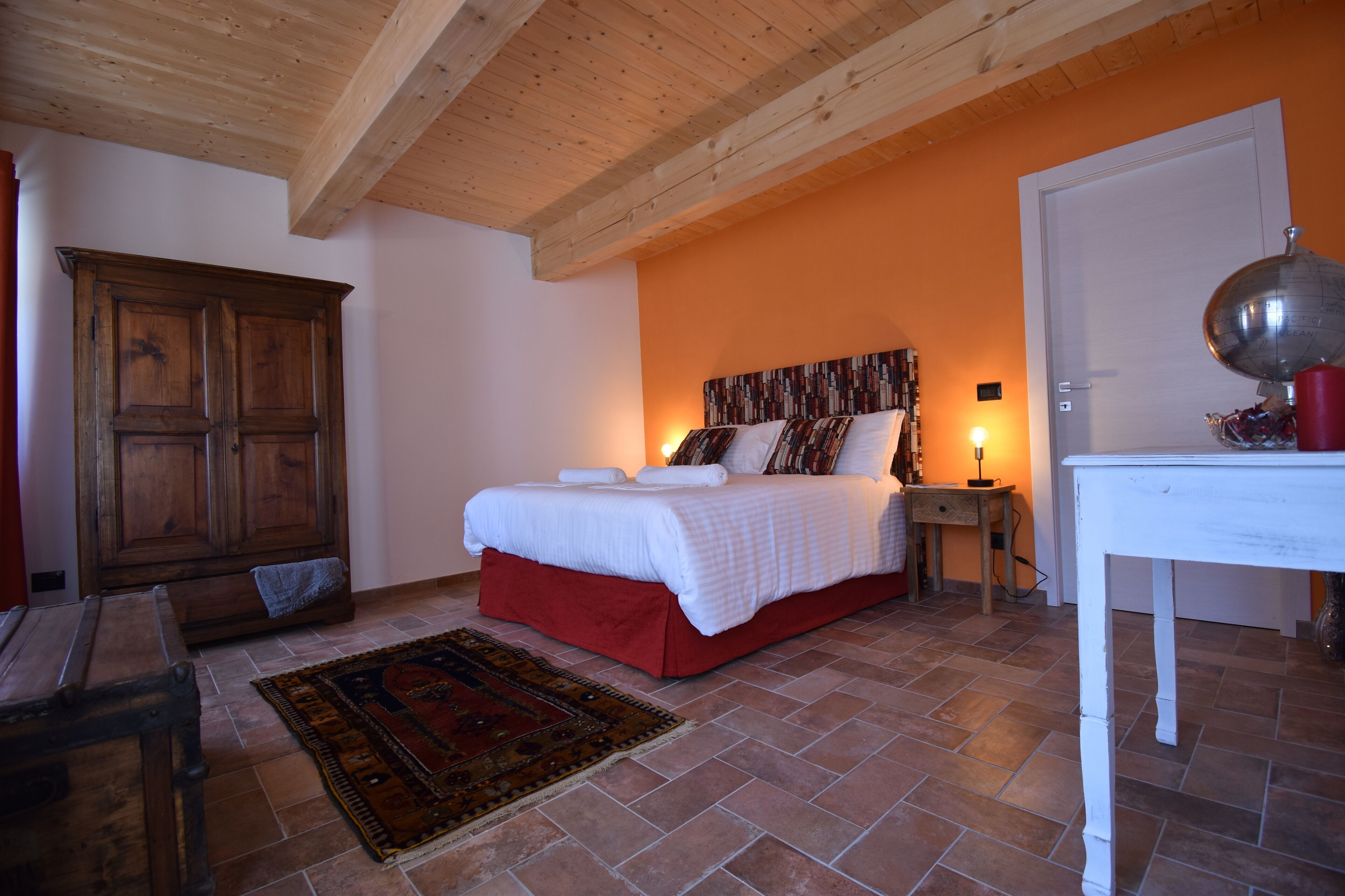 Une chambre avec une armoire en bois, un lit avec une literie blanche, une jupe rouge, une table avec un globe terrestre et une lampe rouge.