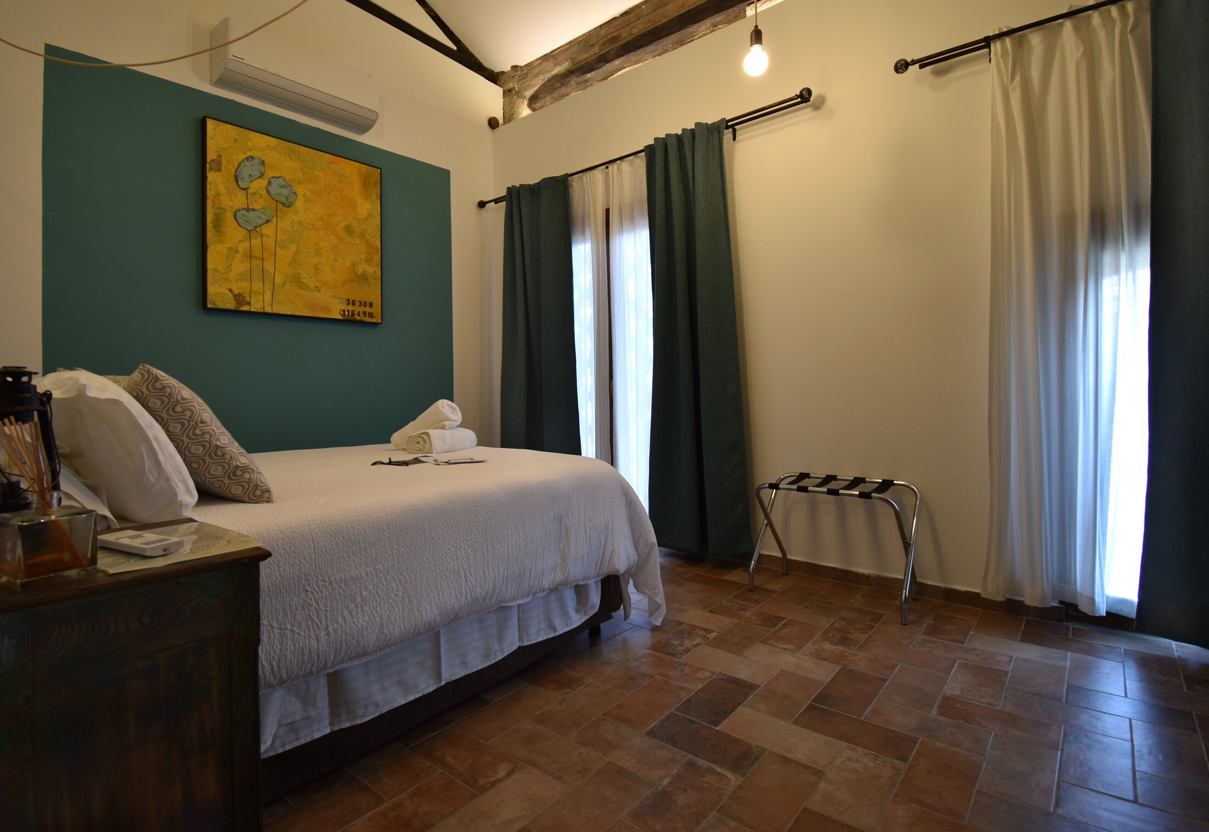A bedroom with a bed, a painting on the wall, a wooden nightstand, and a window with curtains.