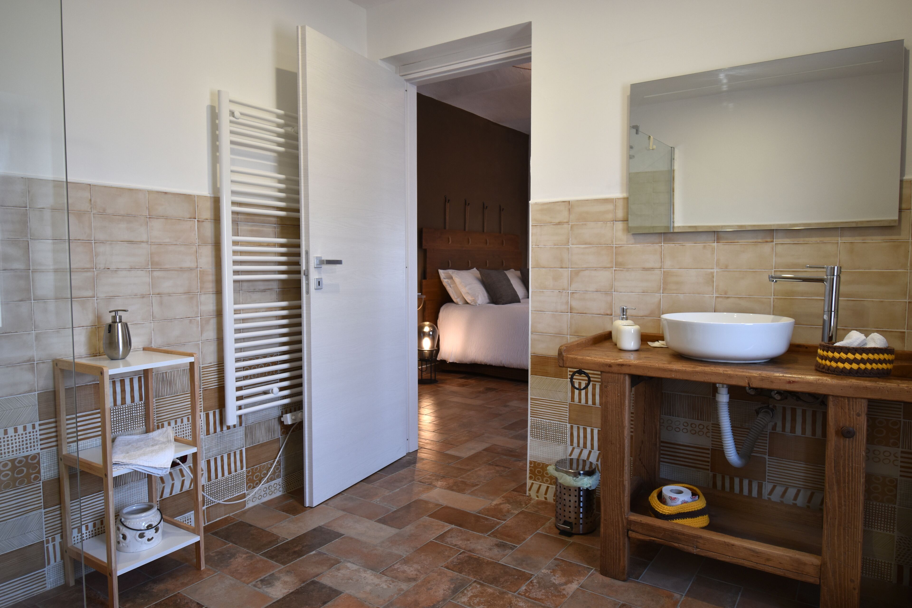 A bathroom with a white sink, mirror, and towel rack. There is a wooden shelf with toiletries and a trash can. The floor is tiled.