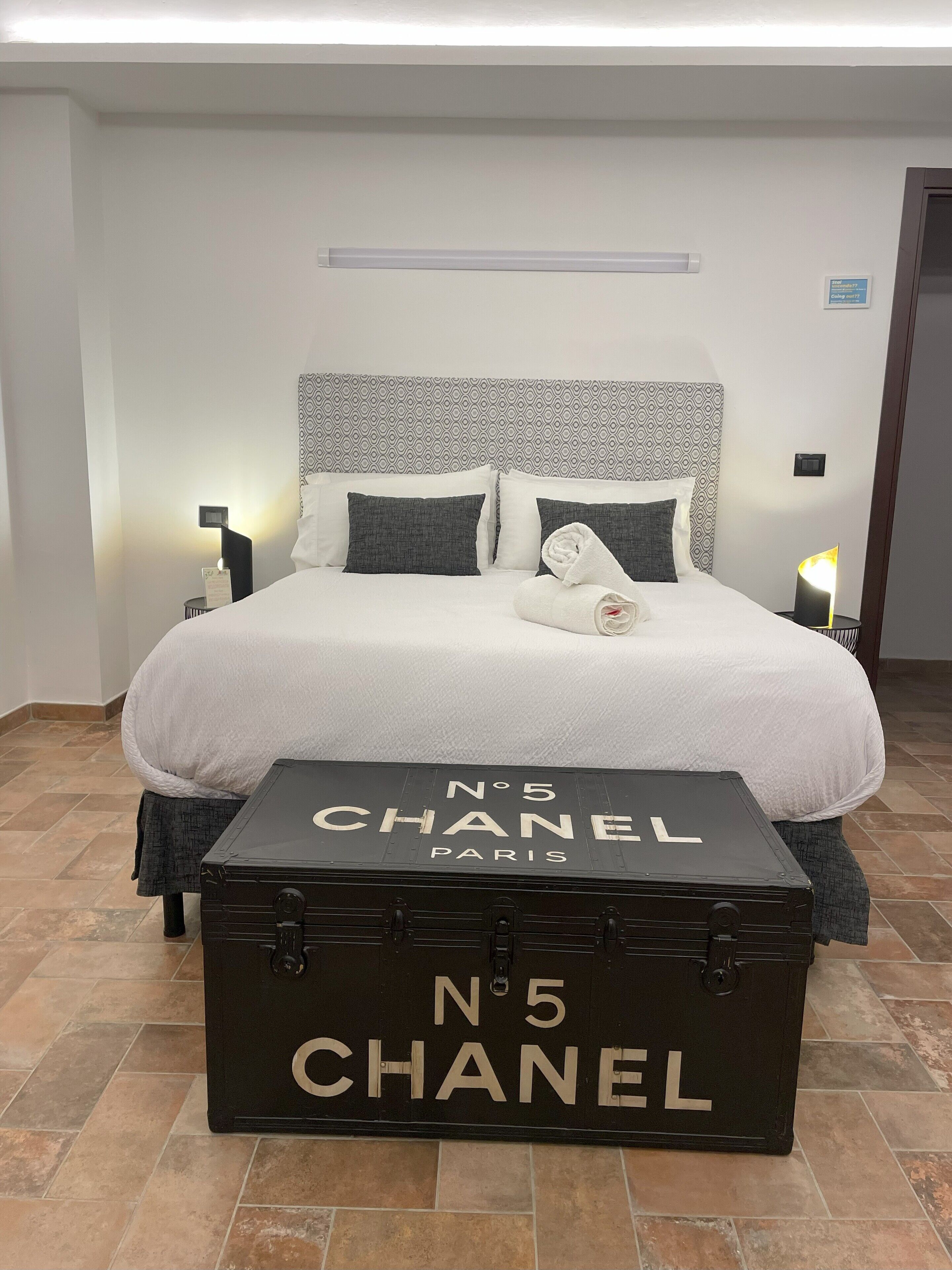 A hotel room with a bed, two bedside tables, a nightstand, a rolled towel, and a Chanel suitcase.