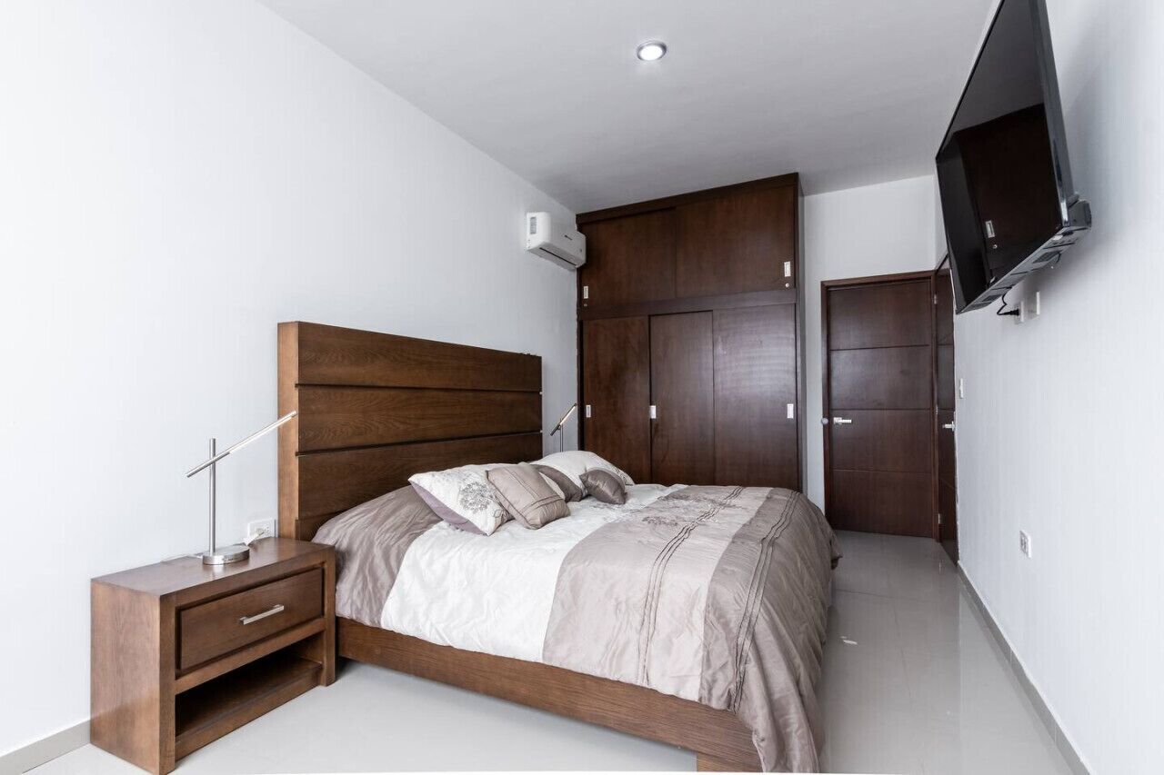 A bedroom with a wooden bed, nightstand, wardrobe, and a mounted TV.