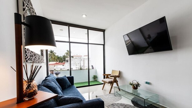 A modern living room with a dark blue sofa, a glass coffee table, a wooden side table with a lamp, a mounted TV, and a view of the outdoors through large windows.