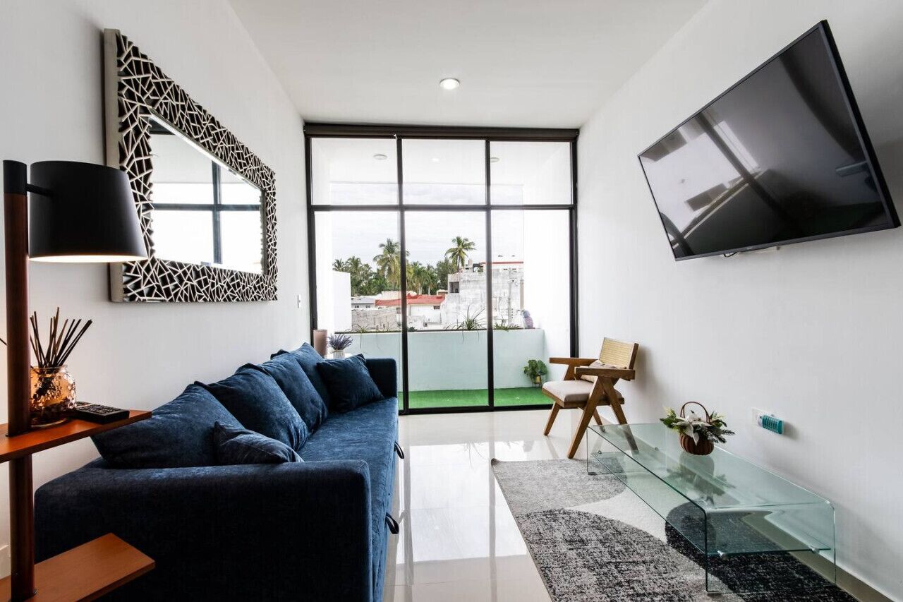 A modern living room with a blue sofa, a glass coffee table, a mounted TV, and a large window with a view of palm trees and buildings.