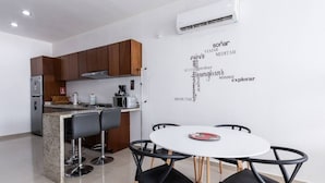 A modern kitchen with a dining area, featuring a round table with four chairs, a built-in island with bar stools, wooden cabinets, and an air conditioning unit on the wall.